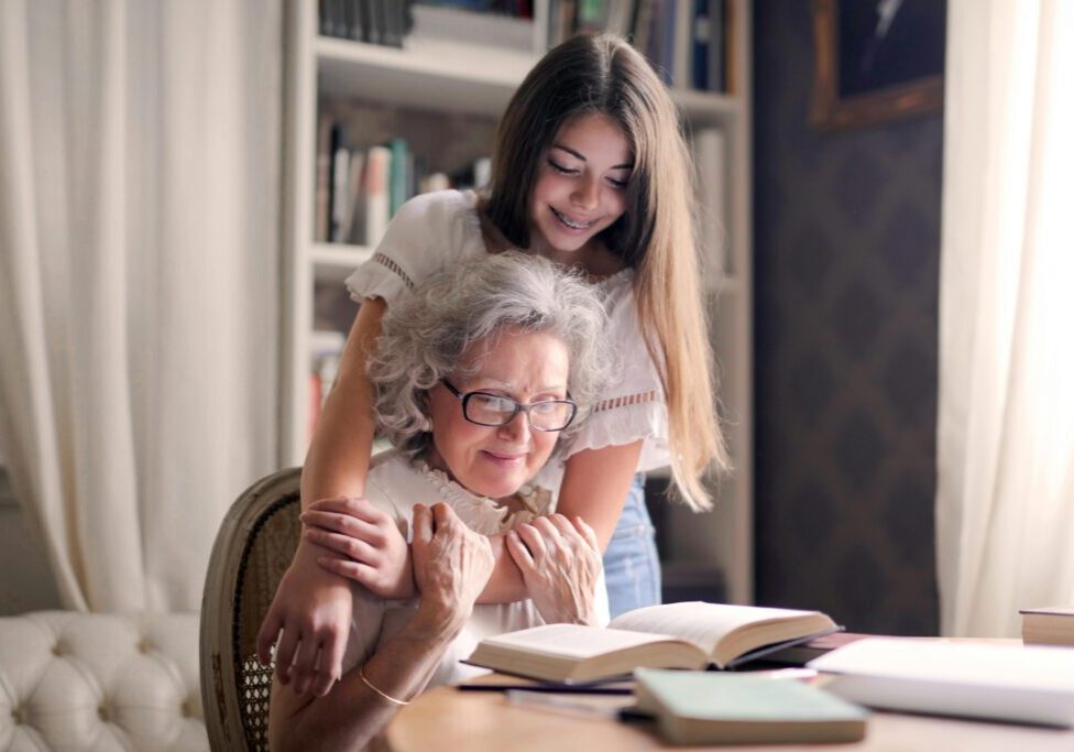 Young woman hugging grandmother reading book.